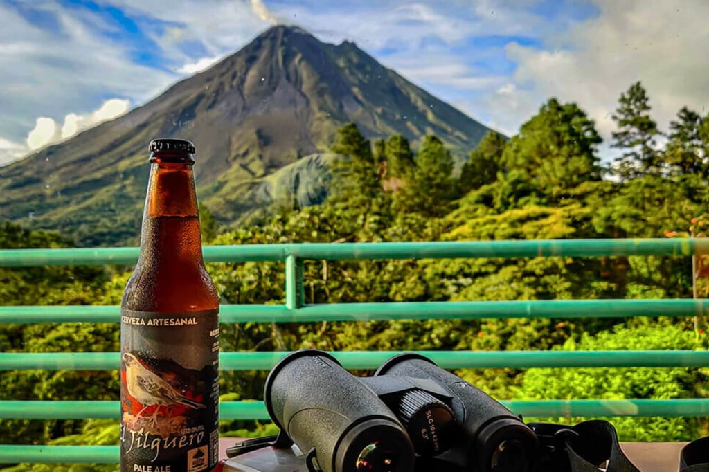 Bierflasche und Fernglas auf Geländer, im Hintergrund ein Vulkan und grüne Vegetation.