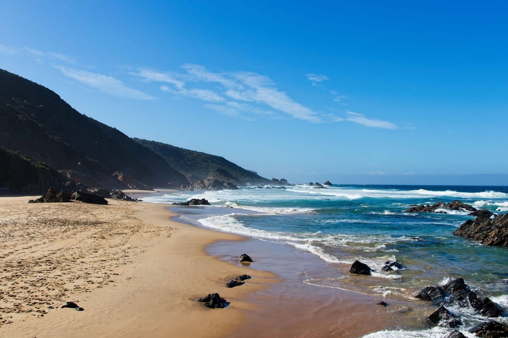 Weitläufiger Sandstrand mit Felsen und Klippen unter blauem Himmel, Wellen schlagen ans Ufer.