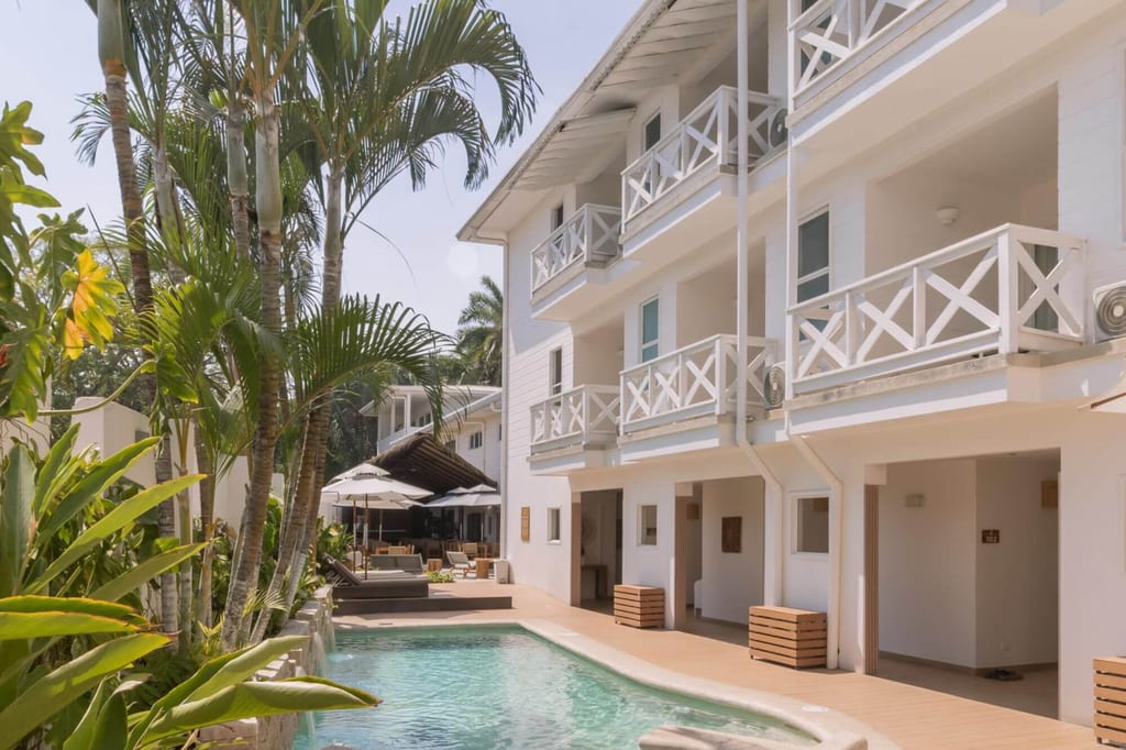 Außenpool neben weißem Resort mit Palmen und Sonnenschirmen bei hellem Tageslicht.