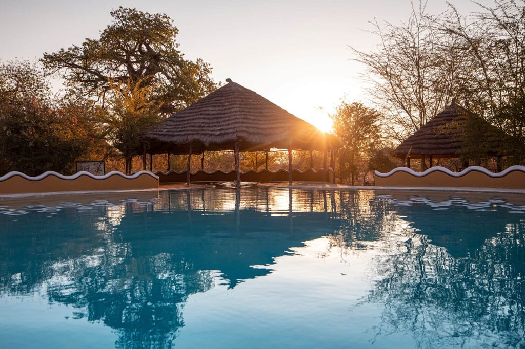 Pool mit spiegelnder Wasserfläche und strohgedeckten Hütten im goldenen Licht tief stehender Sonne.