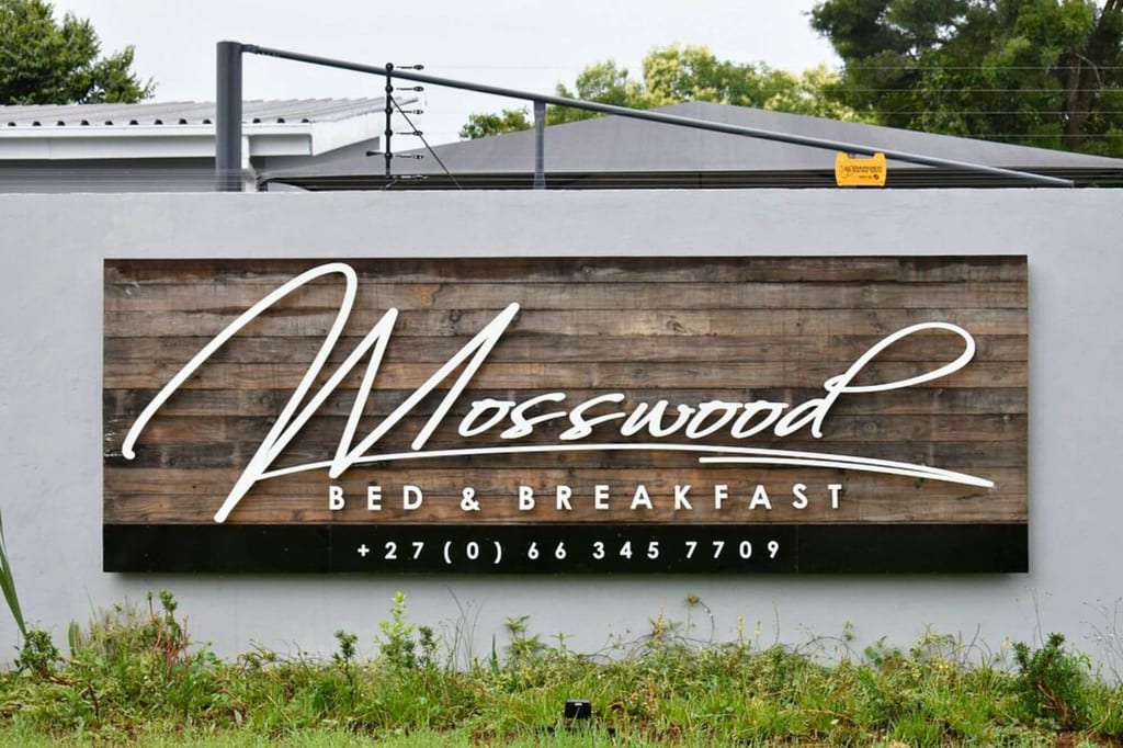 Holzschild mit weißer Aufschrift ‚Mosswood Bed & Breakfast‘ an einer grauen Mauer, Gras im Vordergrund.