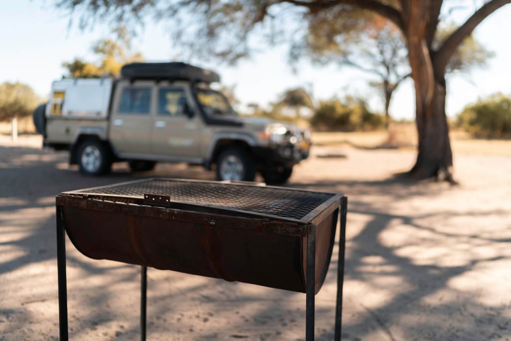 Rostiger Grill im Vordergrund, unscharfer Geländewagen und Bäume bei sonnigem Tageslicht.