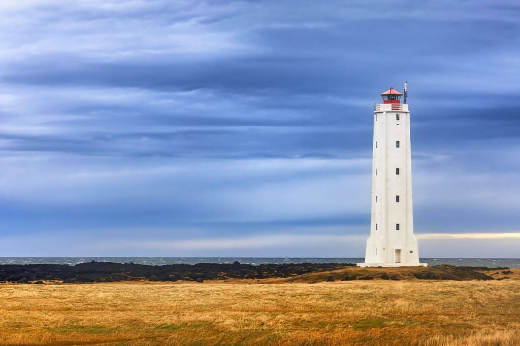Weißer Leuchtturm an zerklüfteter Küste vor dramatischem blau-grauem Himmel, goldene Grasflächen im Vordergrund.