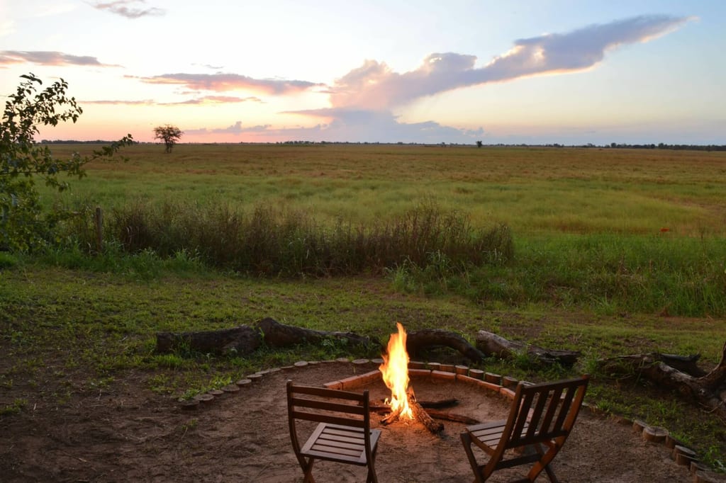 Lagerfeuer mit zwei Holzstühlen vor weiter Graslandschaft bei Sonnenuntergang