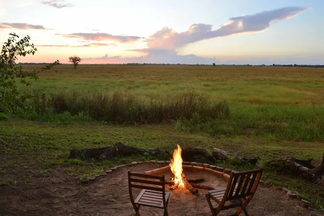 Lagerfeuer mit zwei Holzstühlen vor weiter Graslandschaft bei Sonnenuntergang