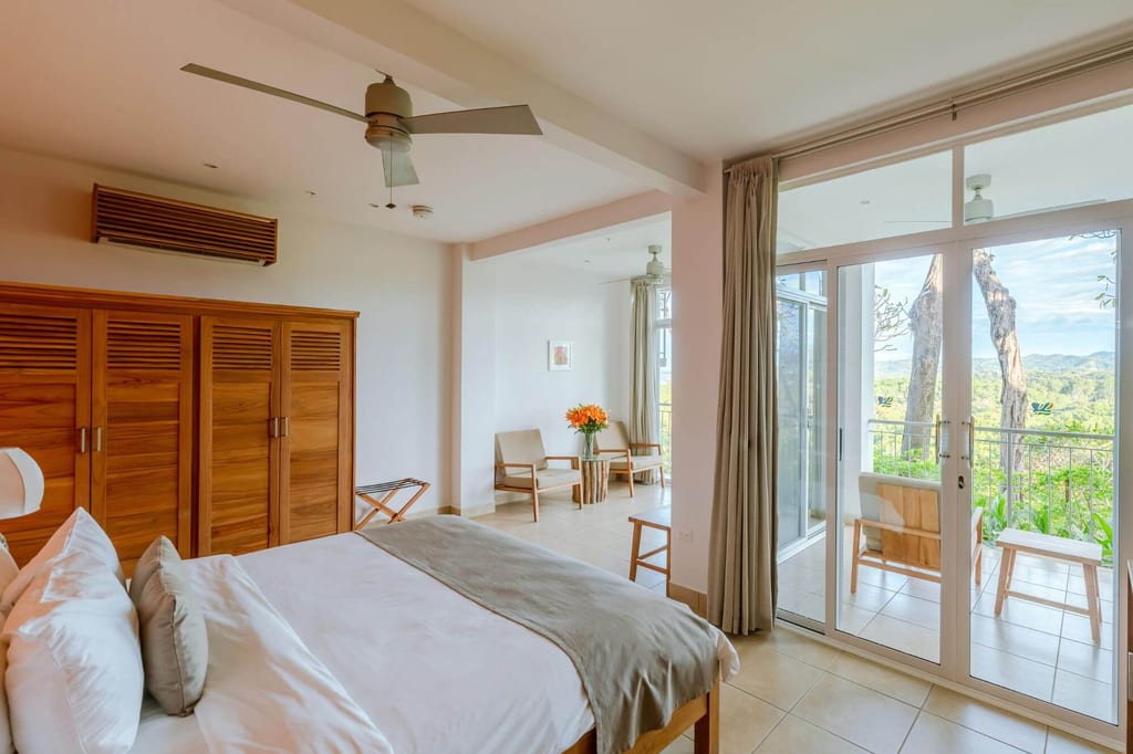 Helles Schlafzimmer mit Doppelbett, Holzmöbeln und Balkon mit Blick auf bewaldete Hügel.