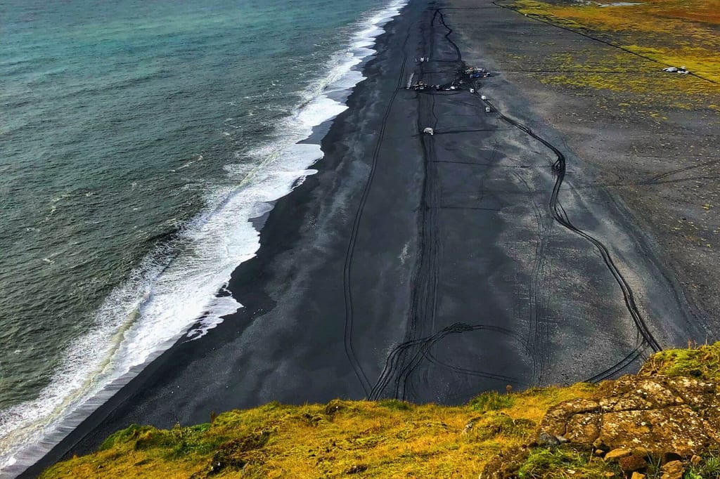 Luftaufnahme eines schwarzen Sandstrands mit Reifen- und Fahrzeugspuren, grasbewachsene Klippe im Vordergrund.