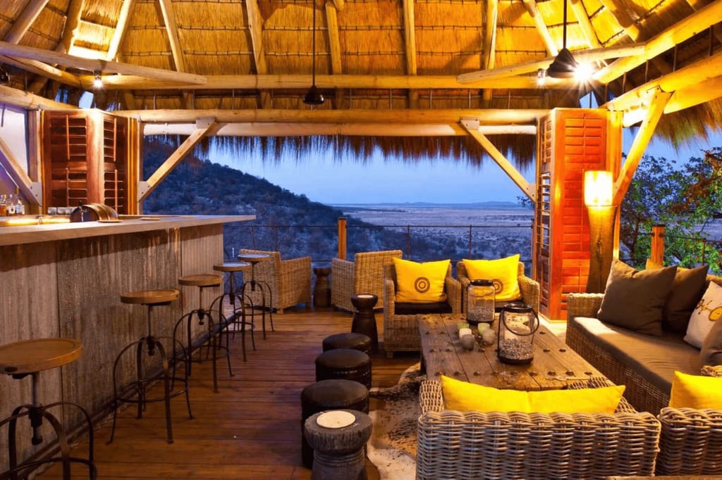 Offene Lodge mit Strohdach und Rattanmöbeln, Blick in die Dämmerung über eine weite Ebene.