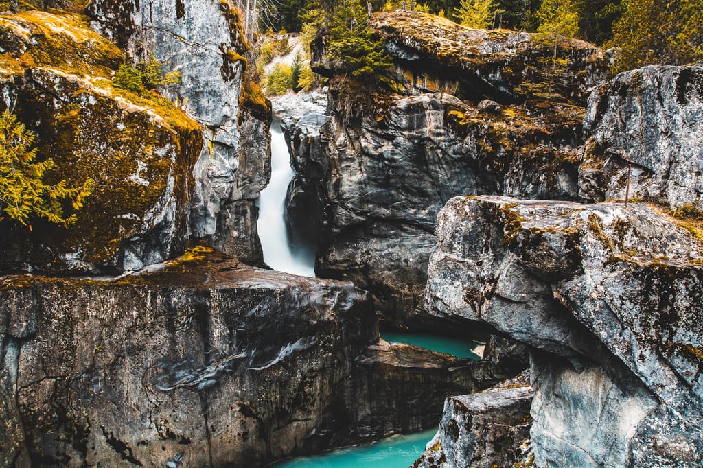 Enges Felsental mit kleinem Wasserfall und türkisfarbenem Becken, moosbedeckte Felsen und Nadelbäume.