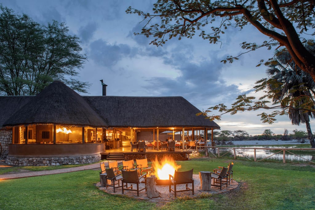 Strohgedeckte Lodge mit Lagerfeuer und Sitzkreis am Seeufer bei Abenddämmerung