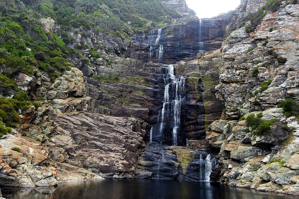 Mehrstufiger Wasserfall auf geschichteten Felsen mit Buschwuchs und dunklem Auffangbecken