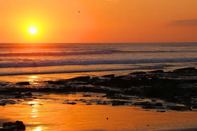 Sonnenuntergang über dem Meer mit Felsen, spiegelnden Reflexionen im Sand und ruhiger Abendstimmung
