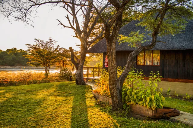 Lodge mit Reetdach am Flussufer im goldenen Abendlicht, Bäume und Rasen im Vordergrund.