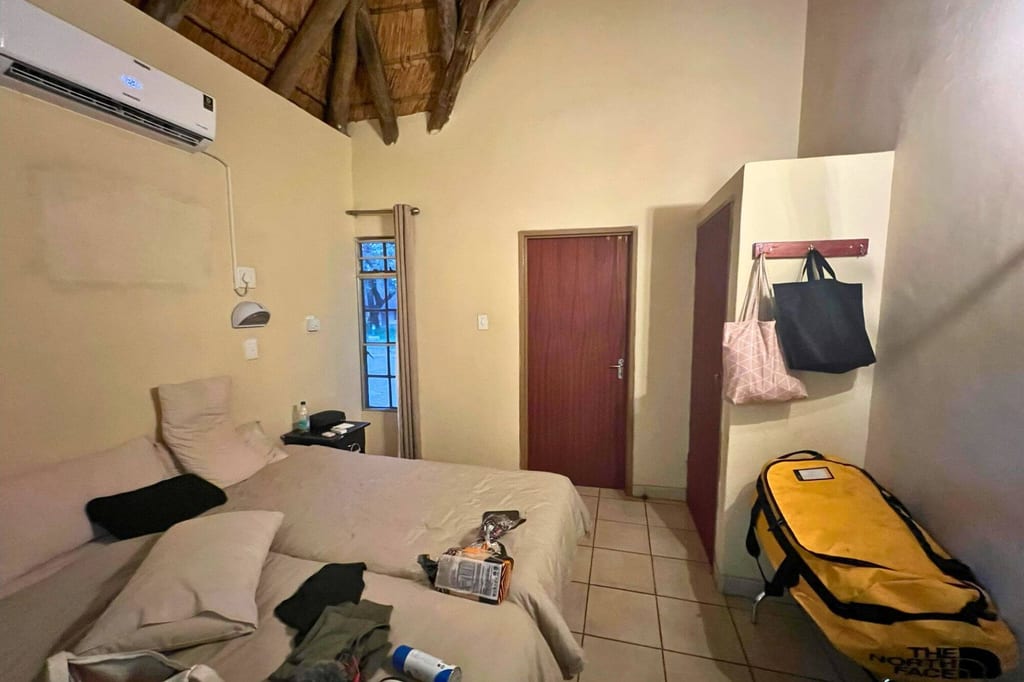 Einfaches Schlafzimmer mit Doppelbett, Strohdach und gelber Reisetasche am Boden.