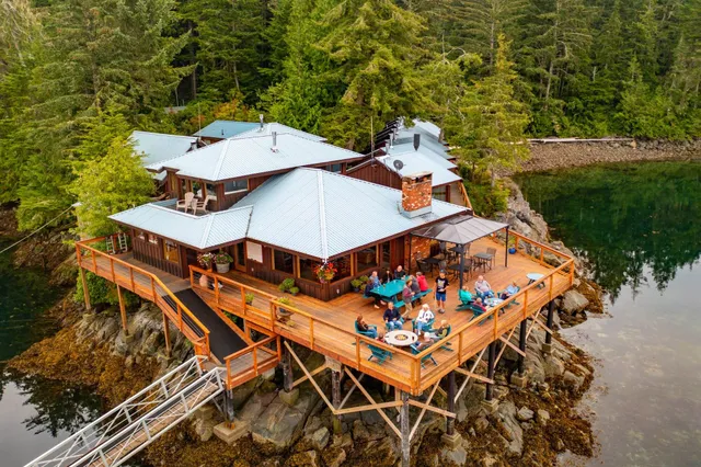 Lodge auf Stelzen mit Menschen auf großem Holzdeck an ruhiger, bewaldeter Küste (Luftaufnahme).