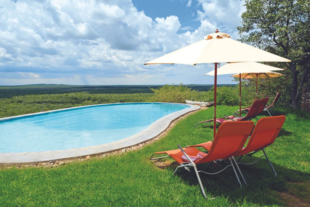 Infinity-Pool mit roten Liegestühlen und Sonnenschirmen vor weiter grüner Landschaft und bewölktem Himmel.