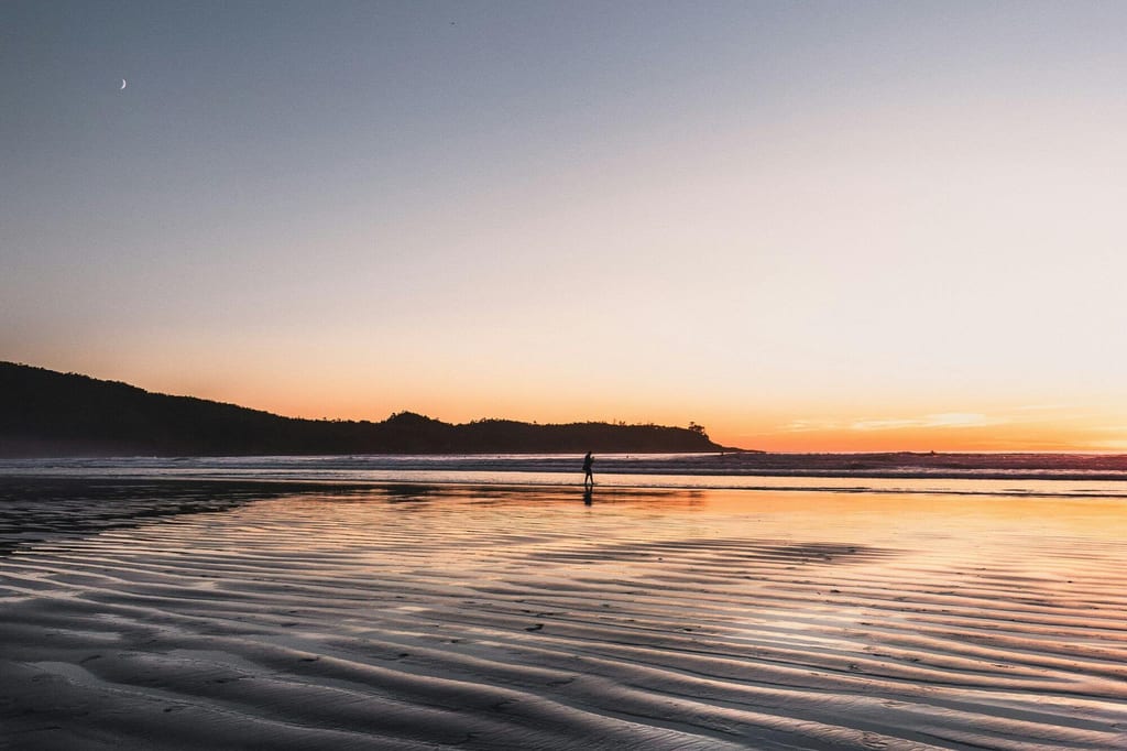 Einsame Person als Silhouette am Strand bei Sonnenuntergang mit nassem, geripptem Sand und Mondsichel.