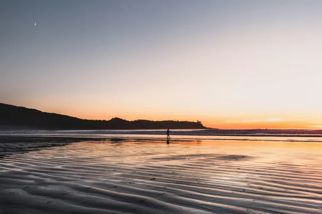Einsame Person als Silhouette am Strand bei Sonnenuntergang mit nassem, geripptem Sand und Mondsichel.