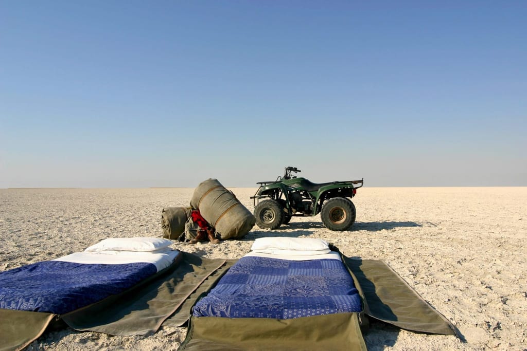 Zwei Feldbetten vor einem ATV und verpackten Bündeln auf einer weiten, flachen Salzlandschaft unter klarem Himmel.