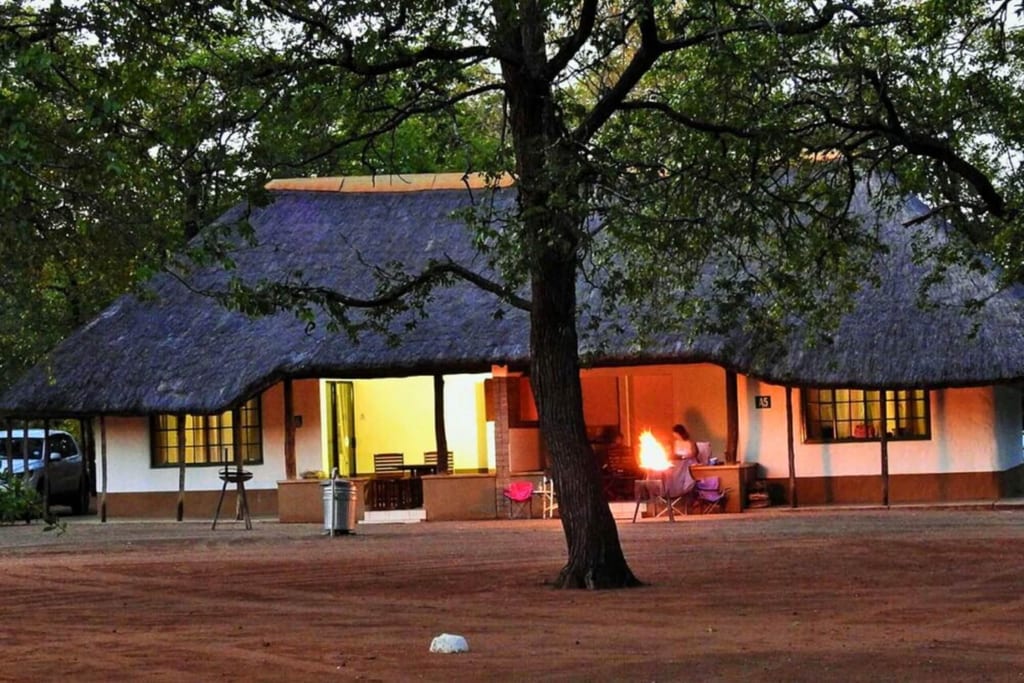 Strohgedeckte Lodge bei Dämmerung, Person sitzt abends am Feuer auf der Veranda