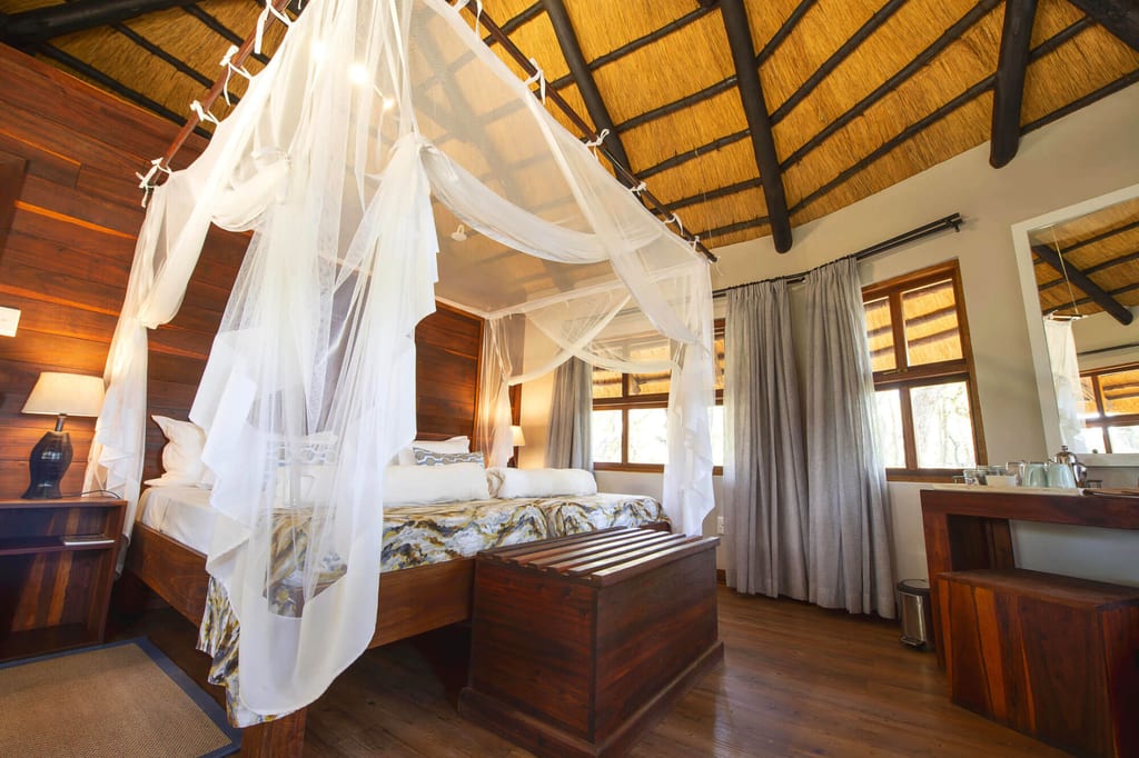 Gemütliches Safari-Schlafzimmer mit Moskitonetz, Strohdach und Holzmöbeln im Tageslicht.