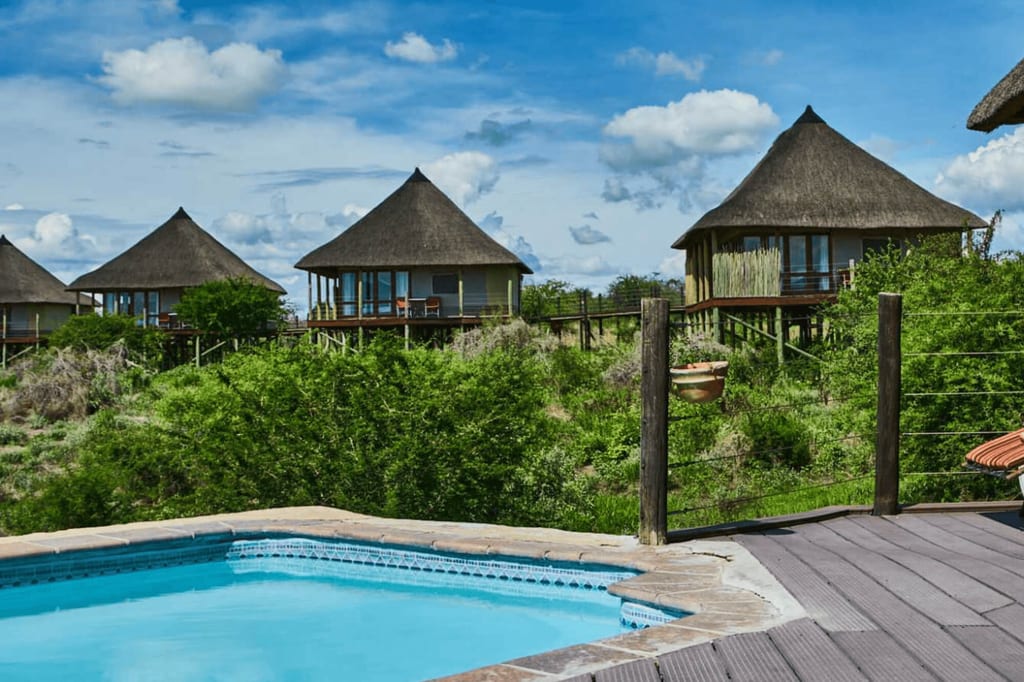 Pool und Holzterrasse im Vordergrund, strohgedeckte Rundhütten auf Stelzen im grünen Buschland