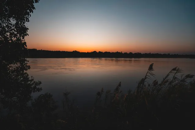 Breiter Fluss bei Sonnenuntergang mit Schilf im Vordergrund und ruhiger Wasseroberfläche