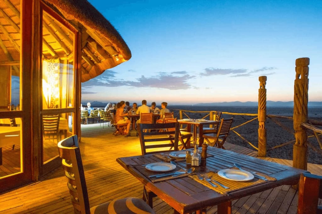 Holzterrasse einer Lodge bei Dämmerung mit gedeckten Tischen und Gästen vor weiter Landschaft.