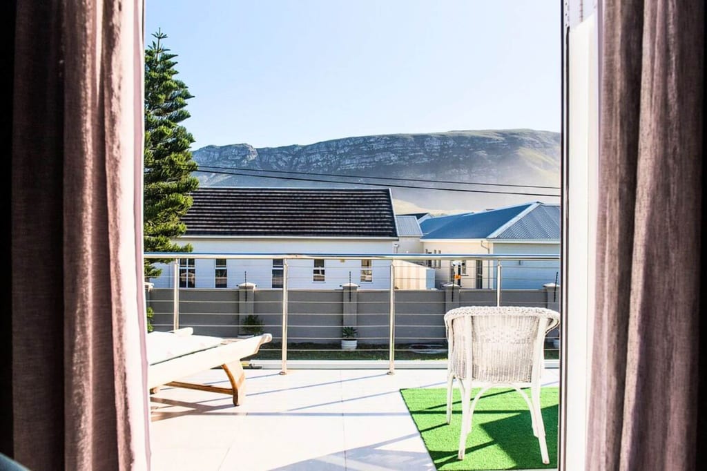 Blick durch Vorhänge auf sonnige Balkonterrasse mit Korbstuhl, Häusern und Berg im Hintergrund