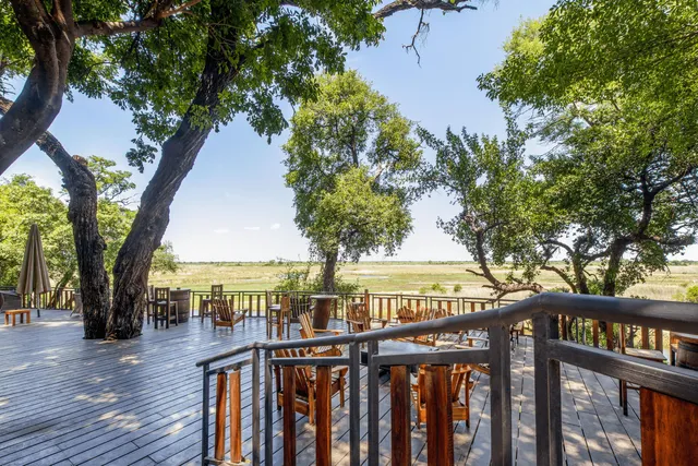 Holzterrasse mit Tischen und Stühlen, Bäume im Vordergrund und weite Graslandschaft im Hintergrund
