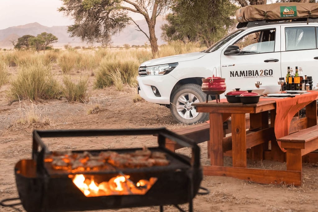Campszene mit Grill im Vordergrund, gedecktem Holztisch und weißem Geländewagen im Abendlicht.