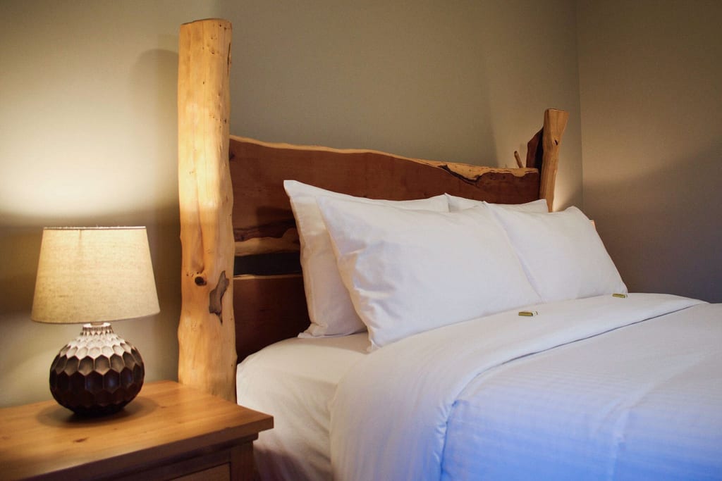 Gemütliches Schlafzimmer mit naturbelassenem Holzkopfteil, weißer Bettwäsche und warm beleuchteter Nachttischlampe.