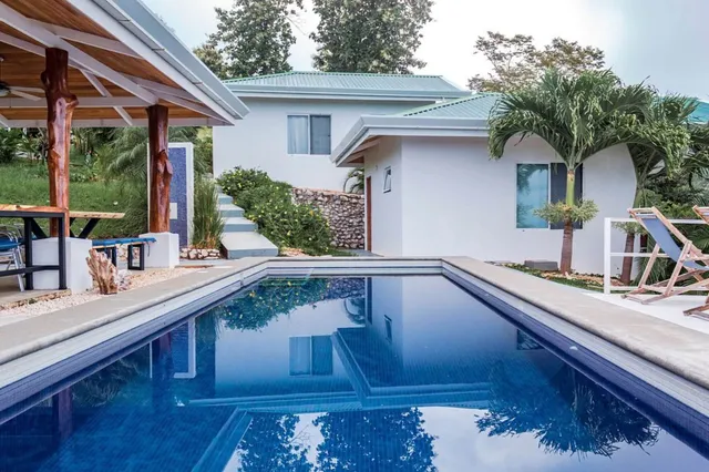 Rechteckiger Pool mit Spiegelung, vor weißen Bungalows und Palmen, ruhige Tageslichtstimmung