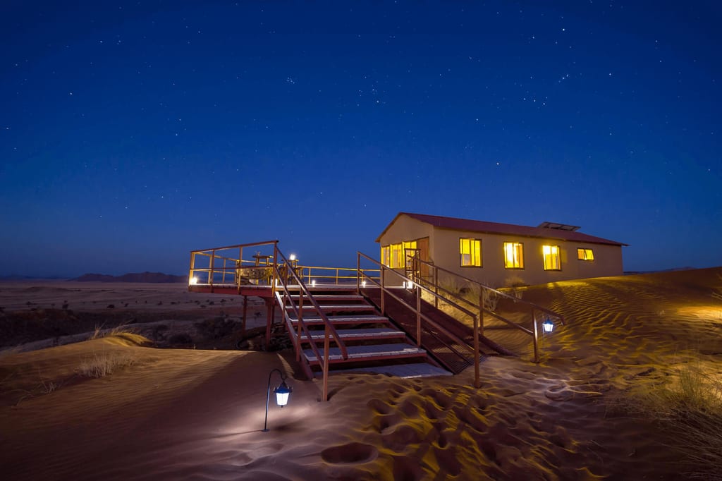 Lodge auf Sanddüne bei Nacht mit beleuchteter Holztreppe und Sternenhimmel