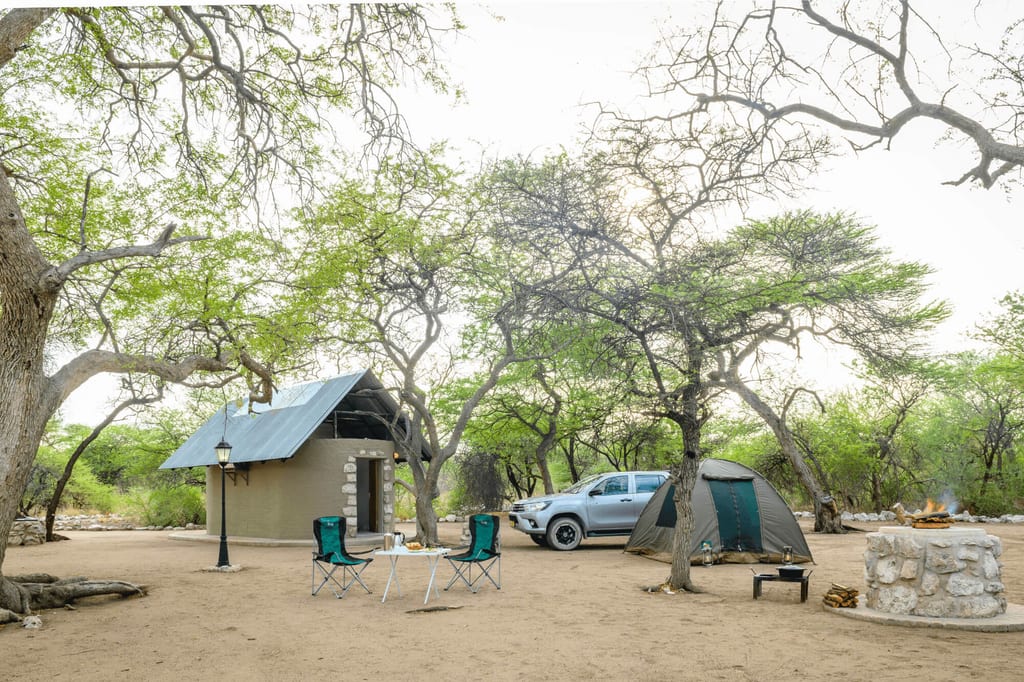 Zwischen Bäumen steht ein Campingtisch mit Stühlen und ein Zelt
