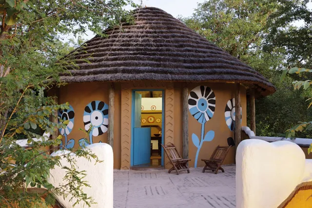 Rundes strohgedecktes Haus mit blauen Blumenmalereien und offener Tür, Terrasse mit Holzstühlen.