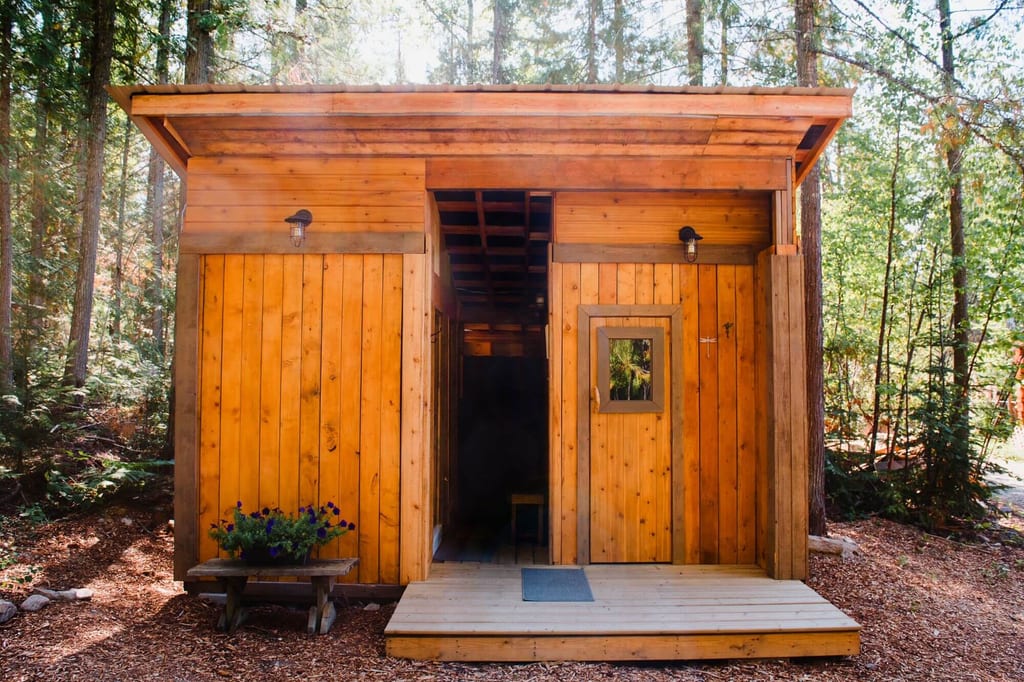 Vorderansicht einer kleinen Holzhütte mit offener Tür und Blumenbank im sonnendurchfluteten Wald.
