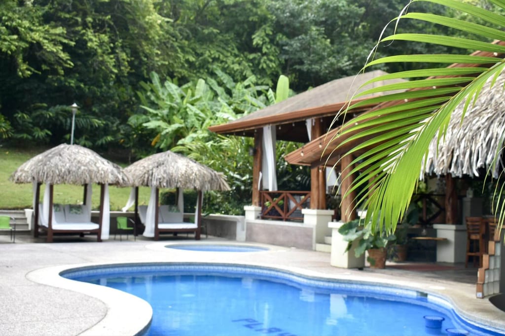 Tropischer Hotelpool mit Strohdach-Cabanas, Holzterrasse und dichter Vegetation bei Tageslicht.