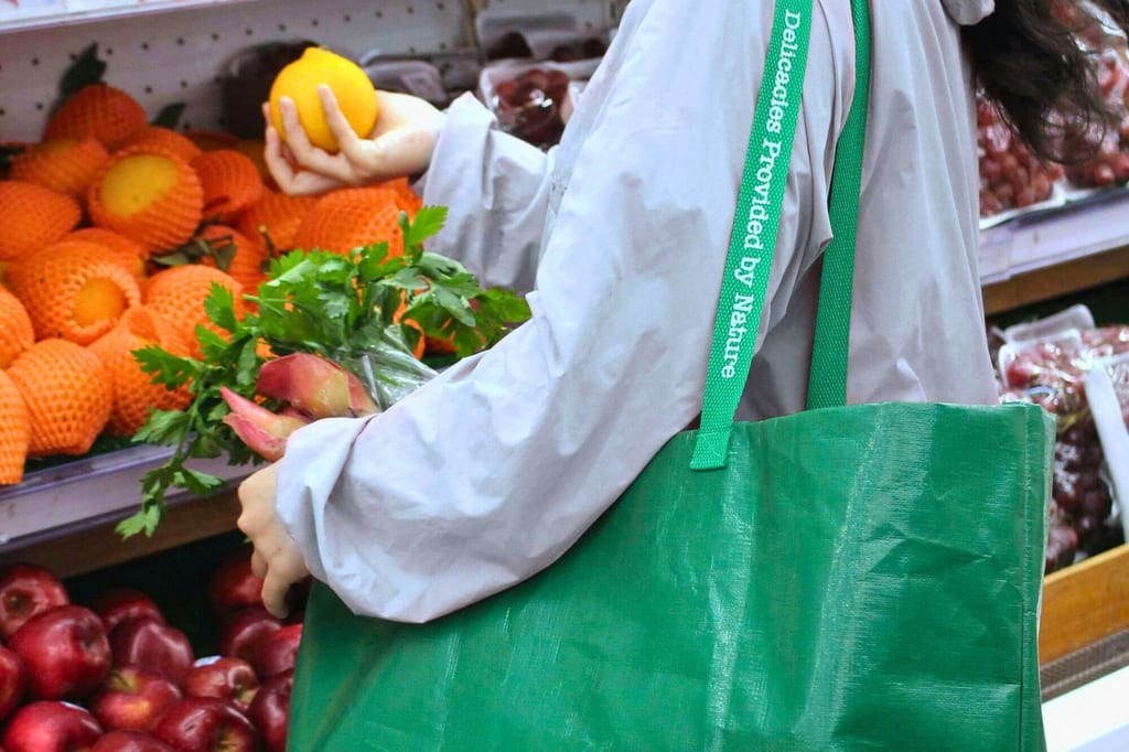 Seitenaufnahme einer einkaufenden Person mit grüner Stofftasche, hält eine Orange vor Obstregalen.