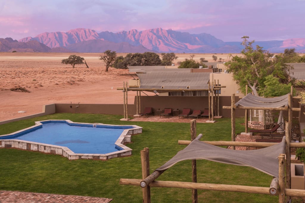 Hotelgarten mit Pool und Pergolen, dahinter Wüste und rosa beleuchtete Berge im Morgen- oder Abendlicht.