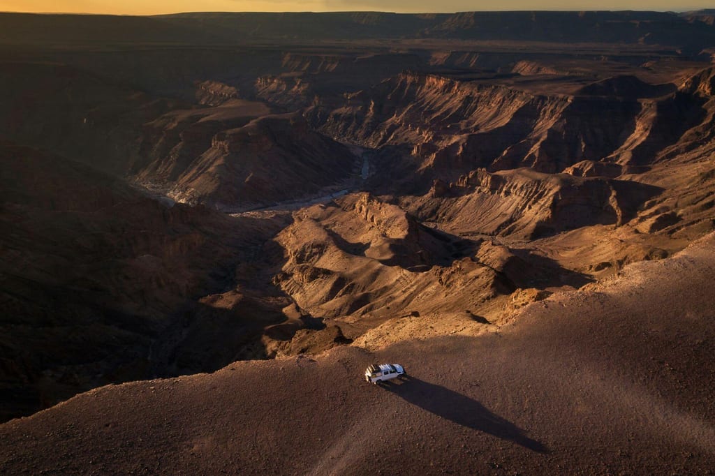 Luftaufnahme eines Geländewagens auf einer Plateaukante über einer weiten, sonnenbeschienenen Schlucht.