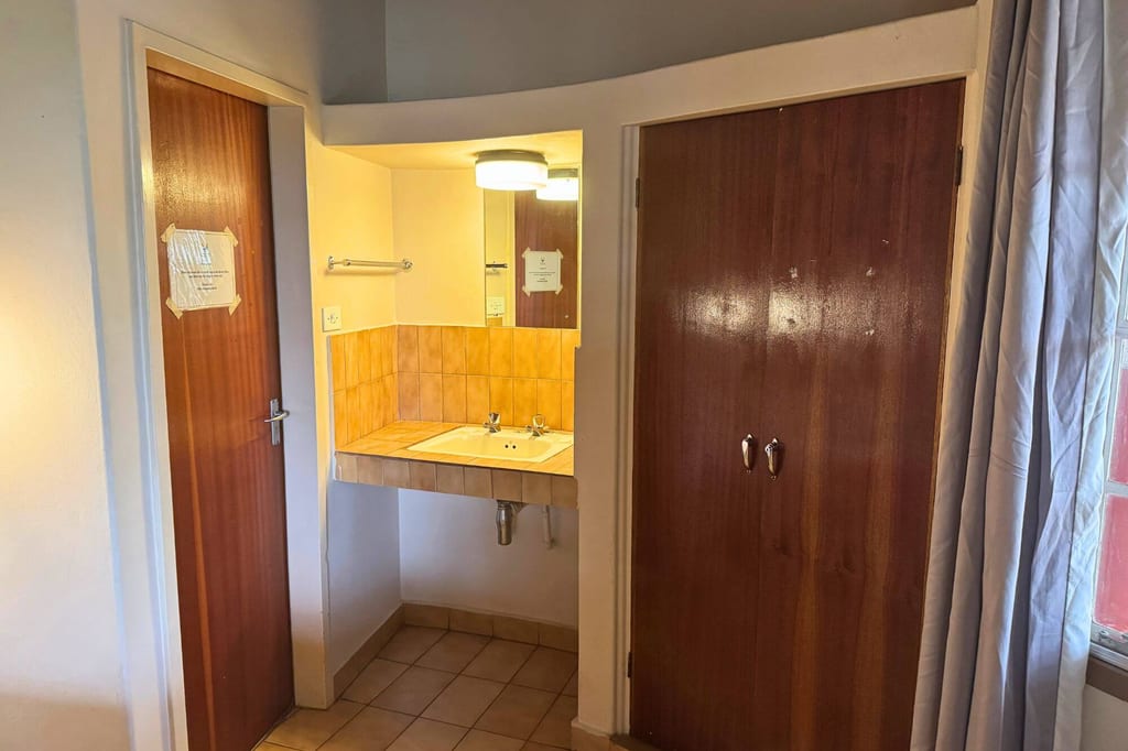 Einfaches Gästezimmer mit Waschbecken in Nische, Holzschranktüren und Vorhang am Fenster.