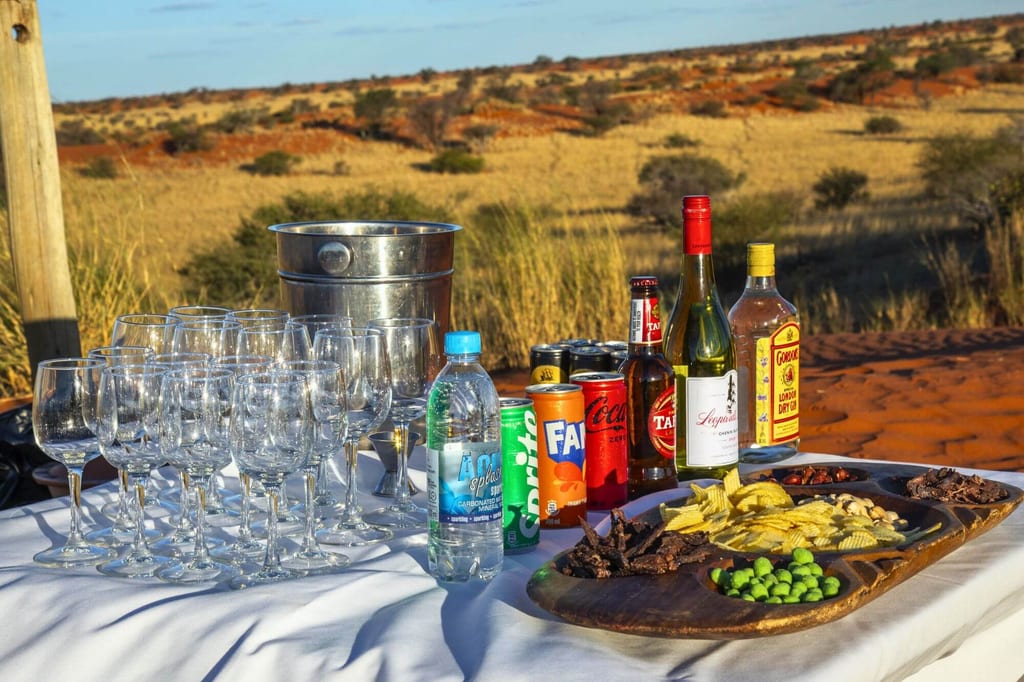 Gedeckter Tisch mit Gläsern, Flaschen, Dosen und Snacks vor trockener Graslandschaft im Abendlicht.