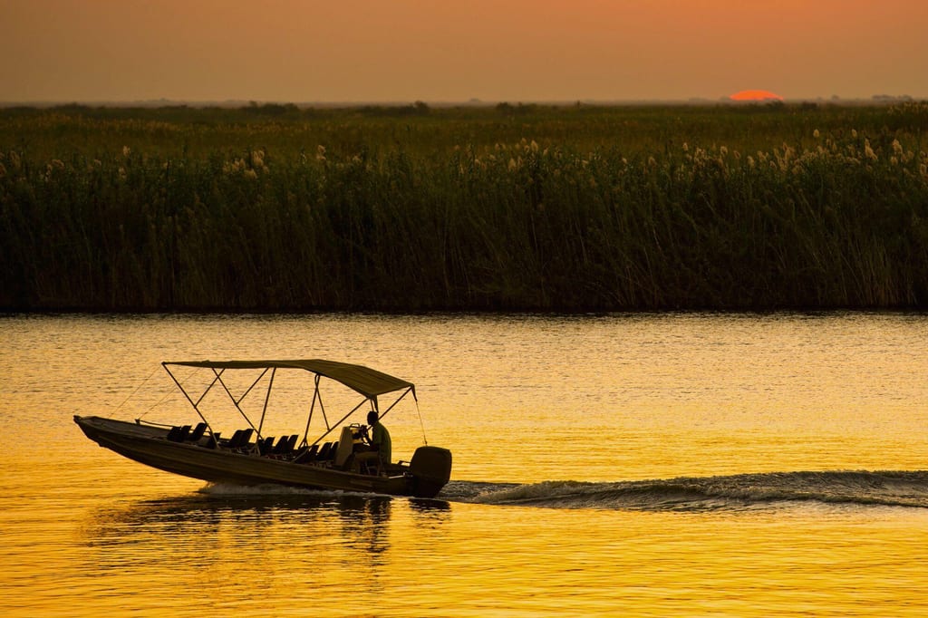 Offenes Motorboot mit Fahrer als Silhouette bei Sonnenuntergang auf einem Fluss in Kasane, Botswana.