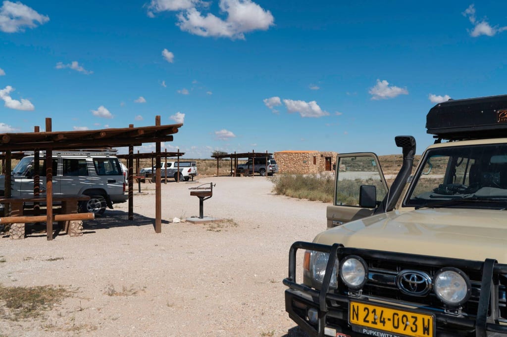 Sonniger Rastplatz mit mehreren Geländewagen, Holzüberdachungen und kleinem steinernen Gebäude