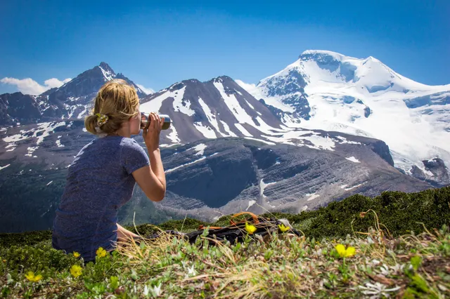Person trinkt auf alpiner Wiese, im Hintergrund schneebedeckte Berggipfel und blauer Himmel.