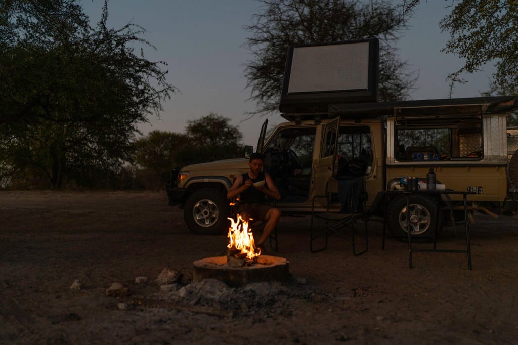 Campingplatz bei Abenddämmerung mit Lagerfeuer, Person und offenem Geländewagen mit Dachzelt.