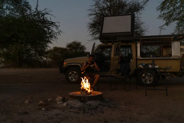 Campingplatz bei Abenddämmerung mit Lagerfeuer, Person und offenem Geländewagen mit Dachzelt.