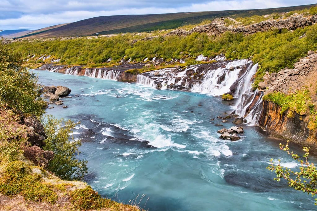 Mehrere Kaskaden eines Wasserfalls in einen türkisfarbenen Fluss, grünes Ufer und bewachsene Hügel im Hintergrund.