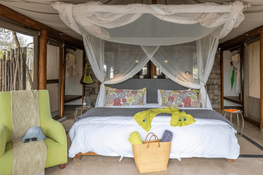 Innenraum eines Safari-Zelts mit Himmelbett, Moskitonetz, grünem Sessel und Korb vor dem Bett.